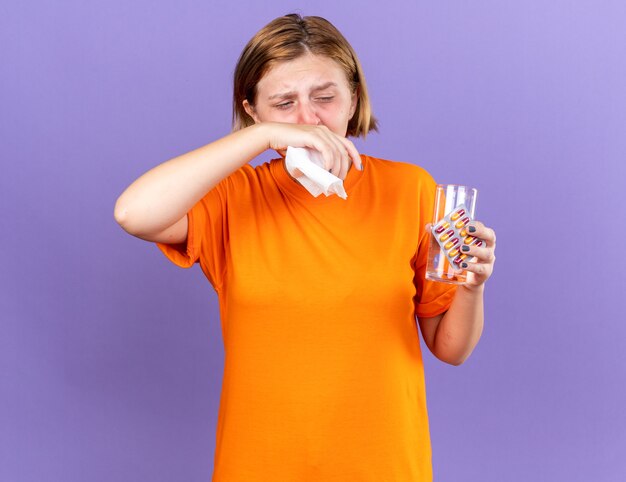 Ungesunde junge Frau in orangefarbenem T-Shirt mit Glas Wasser und Pillen, die sich schrecklich bläst, laufende Nase erwischt hat kaltes Niesen in Taschentuch, das über lila Wand steht