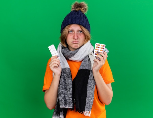 Ungesunde junge Frau in orangefarbenem T-Shirt mit Hut und warmem Schal um den Hals, die schreckliche Pillen hält, die an Viren leiden, die über grüner Wand stehen