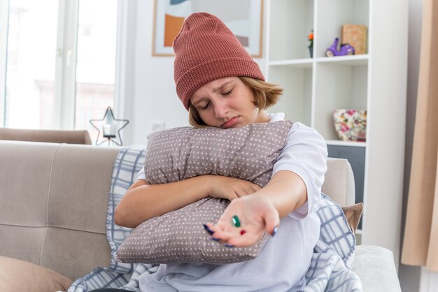 Ungesunde junge Frau in warmem Hut mit Decke, die sich unwohl fühlt und an Erkältung und Grippe leidet, die Kissen und Pillen hält und krank auf der Couch im hellen Wohnzimmer sitzt