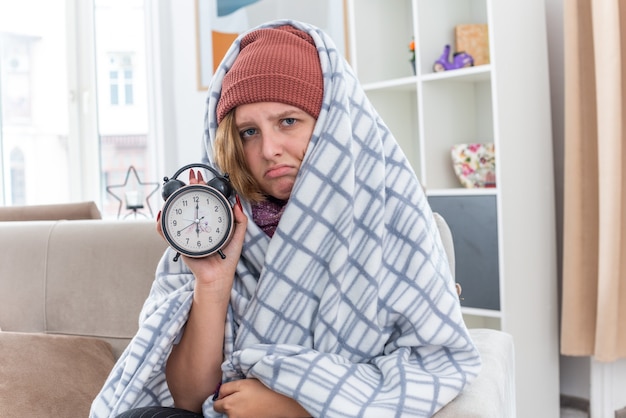 Ungesunde junge Frau mit Hut in Decke gehüllt, die Wecker hält und sich unwohl fühlt und an Erkältung und Grippe leidet und mit traurigem Ausdruck auf der Couch im hellen Wohnzimmer sitzt living