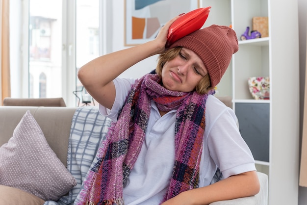 Ungesunde junge Frau mit Hut mit warmem Schal um den Hals, die sich unwohl und krank fühlt und an Erkältung und Grippe leidet, die eine Wärmflasche auf dem Kopf hält und besorgt auf der Couch im hellen Wohnzimmer sitzt