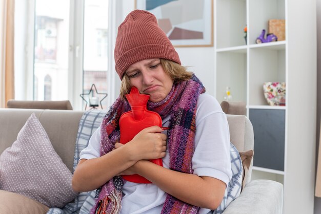 Ungesunde junge Frau mit Hut mit warmem Schal um den Hals, die sich unwohl und krank fühlt und an Erkältung und Grippe leidet, die eine Wärmflasche hält und besorgt auf der Couch im hellen Wohnzimmer sitzt