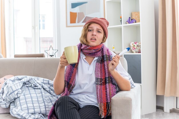 Ungesunde junge Frau mit Hut mit warmem Schal um den Hals, die sich unwohl und krank fühlt und an Erkältung und Grippe leidet, die Thermometer und Tasse besorgt und traurig auf der Couch im hellen Wohnzimmer sitzt living