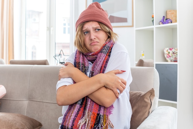 Ungesunde junge Frau mit Hut mit warmem Schal um den Hals, die sich unwohl und krank fühlt und an Erkältung und Grippe leidet und Fieber auf der Couch im hellen Wohnzimmer hat