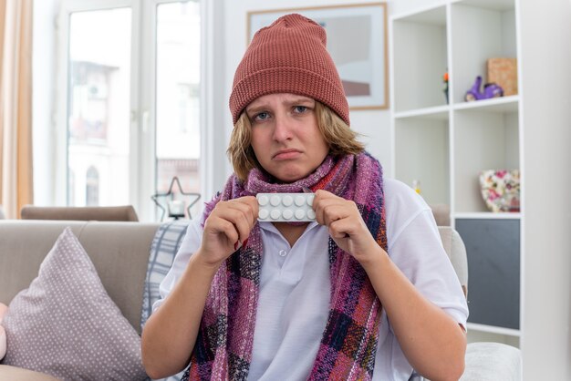 Ungesunde junge Frau mit Hut mit warmem Schal um den Hals, die sich unwohl und krank fühlt und an Erkältung und Grippe leidet und Pillen hält, die besorgt auf der Couch im hellen Wohnzimmer sitzen