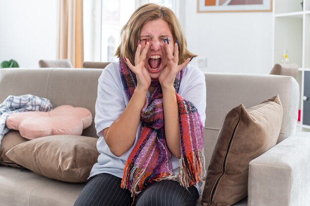 Ungesunde junge Frau mit warmem Schal um den Hals, die schrecklich unter Virusschreien in Panik auf der Couch im hellen Wohnzimmer leidet