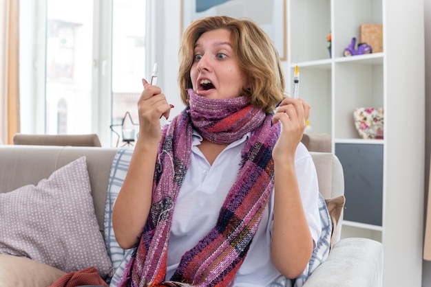 Ungesunde junge frau mit warmem schal um den hals, die sich unwohl und krank fühlt und an erkältung und grippe leidet, die spritze und ampulle hält und besorgt und überrascht aussieht, als sie auf der couch im hellen wohnzimmer sitzt sitting