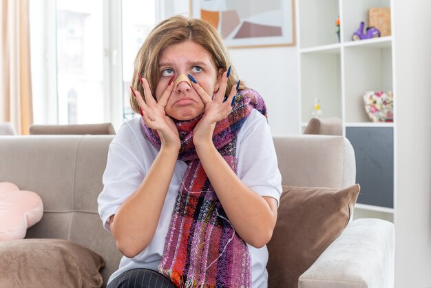 Ungesunde junge Frau mit warmem Schal um den Hals, die sich unwohl und krank fühlt und an Grippe und Erkältung leidet, mit einem Fleck auf ihrer Nase, der verwirrt auf der Couch im hellen Wohnzimmer sitzt