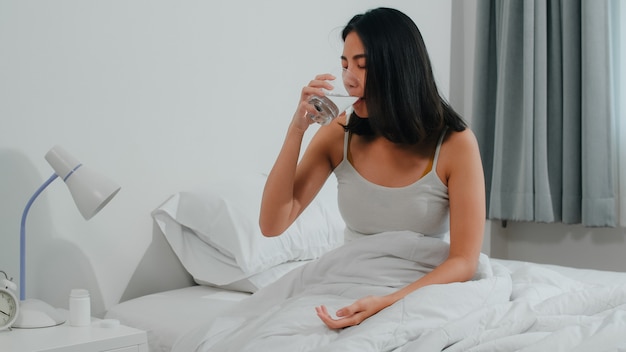 Ungesunde kranke Inderin leidet an Schlaflosigkeit. Asiatische junge Frau, die Schmerzmittelmedizin einnimmt, um die Kopfschmerzenschmerz zu lindern und das Glas Wasser zu trinken, das zu Hause auf Bett in ihrem Schlafzimmer am Morgen sitzt.