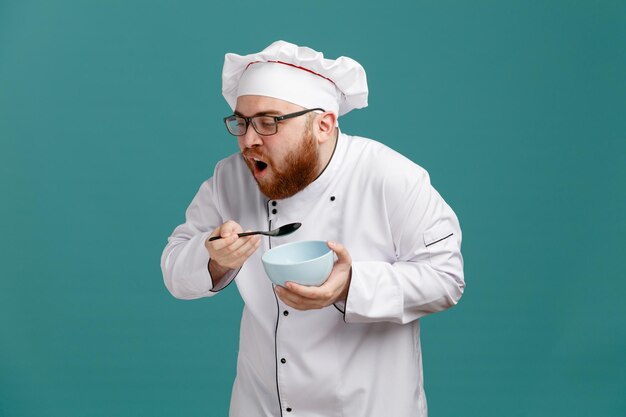 Ungesunder junger männlicher Koch mit Brille, Uniform und Kappe, die eine leere Schüssel und einen Löffel hält, die hustend auf blauem Hintergrund isoliert nach unten schauen