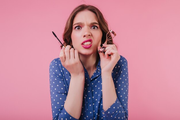 Unglückliche frau mit welligem haar, die ihr make-up vor dem datum macht. nervöses mädchen in blauer kleidung kräuselt wimpern auf rosa wand.