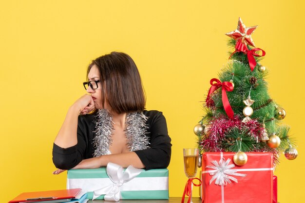 Unglückliche traurige Geschäftsdame im Anzug mit Brille, die ihr Geschenk zeigt und an einem Tisch mit einem Weihnachtsbaum darauf im Büro sitzt