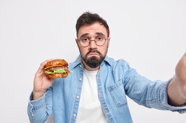 Unglücklicher bärtiger erwachsener europäischer Mann isst Müll hält leckeren Hamburger macht Selfie-Geldbörsen Lippen hat Unzufriedenheit Gesichtsausdruck trägt runde Brille Jeanshemd