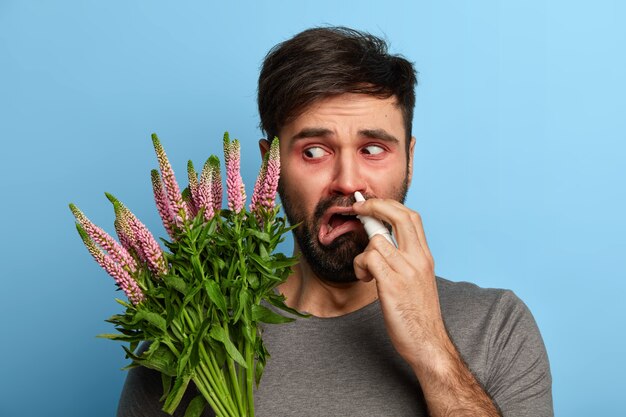 Unglücklicher Mann fühlt sich unwohl, ist allergisch gegen Pollen, leidet an einer Allergie gegen Pflanzen, verwendet Nasenspray für die Nase, braucht medizinische Behandlung, posiert über der blauen Wand, heilt Rhinitis. Medizinisches Konzept.