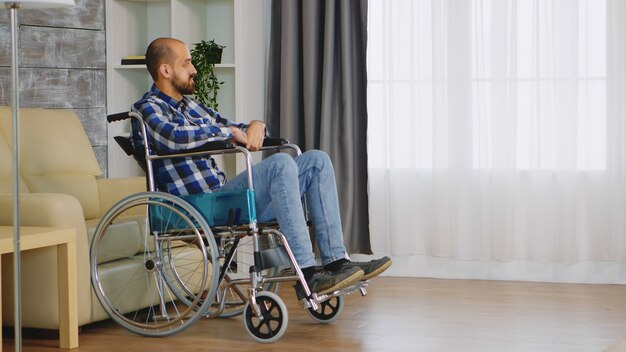 Unglücklicher Mann im Rollstuhl im Wohnzimmer mit Blick auf das Fenster.