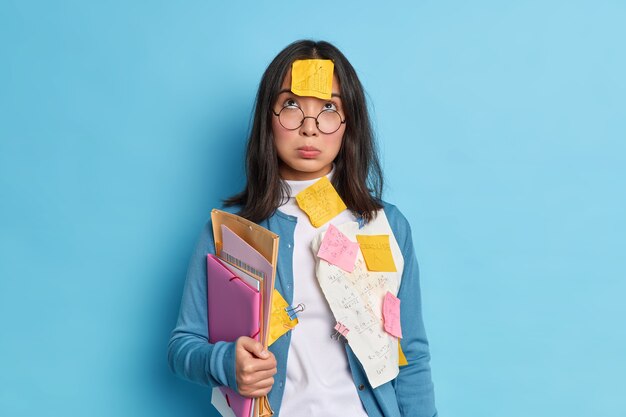 Unglücklicher müder asiatischer Student, der oben konzentriert ist, hat traurigen Ausdruck, trägt eine Brille zur Sehkorrektur hält Ordnerpapiere, die mit Büroklammern geklebt werden.