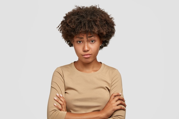 Unglückliches junges weibliches Model mit buschigem Afro-Haarschnitt hat den Gesichtsausdruck missfallen, drückt die Daumen, hört negative Nachrichten vom Gesprächspartner, trägt einen lässigen beigen Pullover und steht alleine im Haus