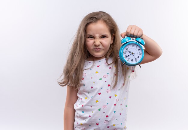 Unglückliches kleines Schulmädchen, das weißes T-Shirt hält Wecker auf isolierter weißer Wand hält