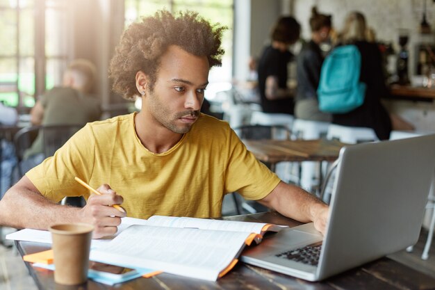 Universitätsstudent mit dunkler Hautfarbe und afrikanischer Frisur, der im Café sitzt und mit Büchern und Notizbuch arbeitet, während er sich auf die Prüfung vorbereitet, die notwendigen Informationen im Internet findet, die ernstes Aussehen haben