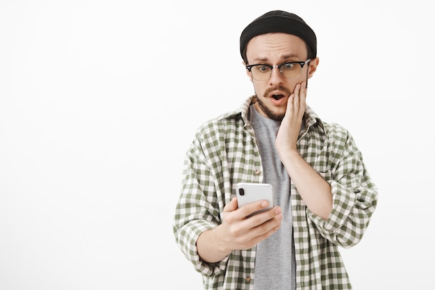 Unpopulärer Kerl, der schockiert ist, Likes unter dem letzten Beitrag im sozialen Netzwerk zu erhalten, der mit geöffnetem Mund nach Luft schnappt und Hand auf Wange hält, überrascht und erstaunt auf Smartphonebildschirm über weißer Wand starrt