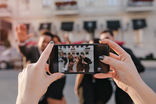 Unscharfes Außenporträt von Frauen und Jungen, die vor dem Gebäude vor der Party mit dem Smartphone im Fokus aufwerfen