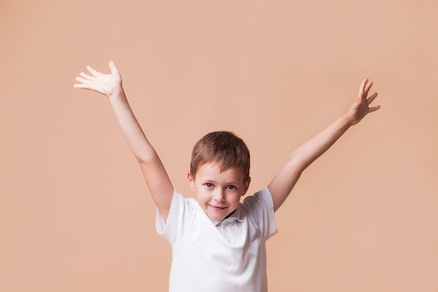 Unschuldiger lächelnder Junge mit der Hand hob Stellung vor beige Hintergrund an