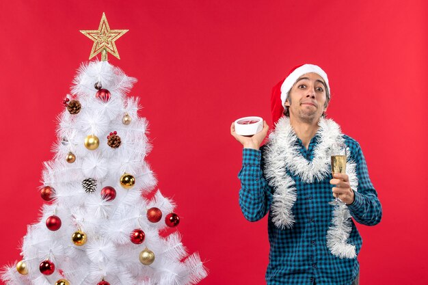 Unsicher verwirrter junger Mann mit Weihnachtsmannhut, der ein Glas Wein hebt und die Uhr stehend hält