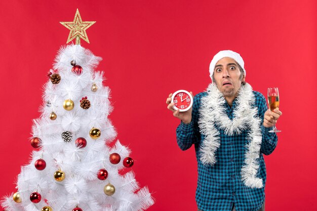 Unsicherer junger Mann mit Weihnachtsmannhut und hält ein Glas Wein und eine Uhr in der Nähe des Weihnachtsbaumes