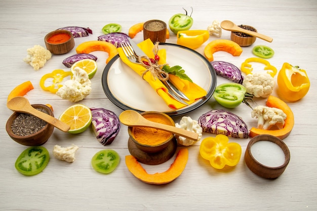 Unten Ansicht gebundenes Messer und Gabel mit gelber Serviette auf runder Platte geschnittenes Gemüse Rotkohlkürbis Blumenkohl gelbe Paprika Gewürze in kleinen Schalen auf weißem Holztisch