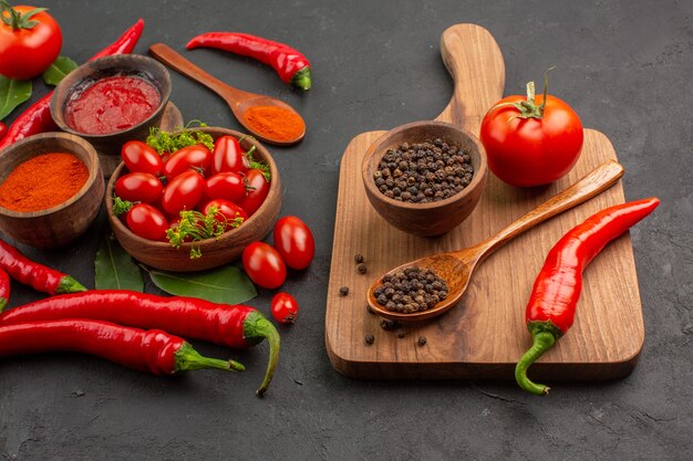 Unten sehen Sie eine Schüssel mit Kirschtomaten, heißen roten Paprikaschoten, Lorbeerblättern und einer Schüssel mit schwarzem Pfeffer, einem Holzlöffel, einer Tomate und einem roten Pfeffer auf dem Schneidebrett auf schwarzem Grund