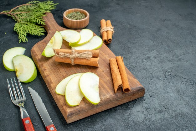 Unteransicht Apfelscheiben und Zimtstangen auf Holzbrettmesser und Gabel getrocknetes Minzpulver in kleiner Schüssel auf dunklem Tisch mit freiem Platz