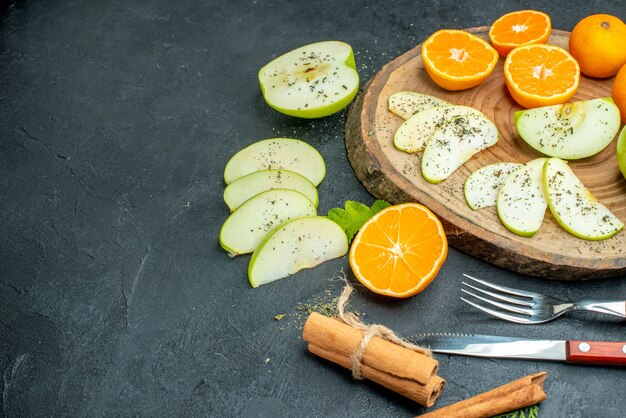 Unteransicht, die Äpfel und Mandarinen mit getrocknetem Minzpulver auf Holzbrett-Zimtgabel und -messer auf dunklem Tisch hackt