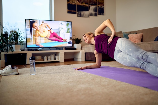 Kostenloses Foto unteransicht einer glücklichen athletin, die in ihrem wohnzimmer in seitlicher plank-pose trainiert