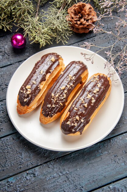 Untergrundansicht Schokoladen-Eclairs auf weißem ovalem Teller-Tannenbaumzweigkegel-Weihnachtsspielzeug auf dunklem Holztisch