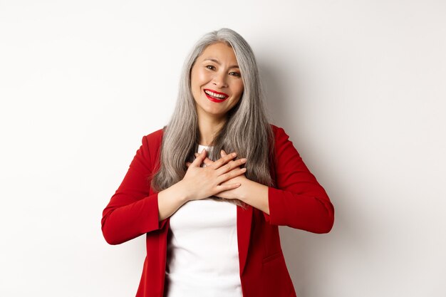 Unternehmenskonzept. Reife asiatische Frau mit roten Lippen und Blazer, Händchen haltend am Herzen und dankbar lächelnd, dankbar in die Kamera schauend, über weißem Hintergrund stehend.