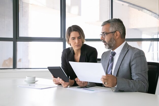 Unternehmensleiter analysieren Berichte. Zwei Geschäftskollegen sitzen zusammen, betrachten Dokument, halten Tablette und sprechen. Mittlerer Schuss. Kommunikationskonzept