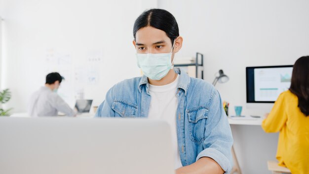 Unternehmer aus Asien, der eine medizinische Gesichtsmaske für soziale Distanzierung in einer neuen normalen Situation zur Virenprävention trägt, während Laptop wieder bei der Arbeit im Büro verwendet wird. Lebensstil nach dem Koronavirus.