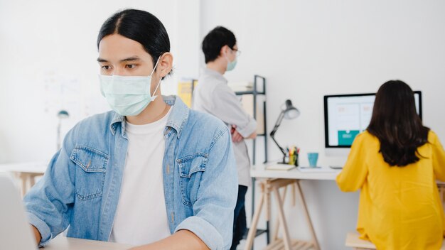 Unternehmer aus Asien, der eine medizinische Gesichtsmaske für soziale Distanzierung in einer neuen normalen Situation zur Virenprävention trägt, während Laptop wieder bei der Arbeit im Büro verwendet wird. Lebensstil nach dem Koronavirus.