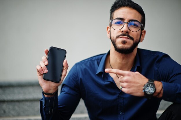 Unternehmer aus dem Nahen Osten tragen eine Brille mit blauem Hemd gegen ein Bürogebäude, das auf der Treppe sitzt, und zeigen den Finger zum Telefon