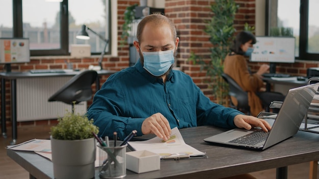 Unternehmer, die auf den Bildschirm schauen und mit einem Laptop arbeiten, um Tarifdiagramme auf Papieren für die Projektplanung am Schreibtisch zu entwerfen. Mann, der Computer verwendet, um Marketingstrategie zu planen und Gesichtsmaske zu tragen.