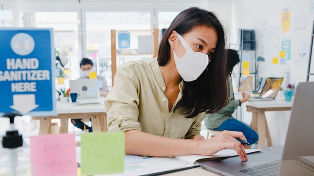 Unternehmerin aus Asien, die eine medizinische Gesichtsmaske für soziale Distanzierung in einer neuen normalen Situation zur Virenprävention trägt, während sie den Laptop wieder bei der Arbeit im Büro verwendet. Leben und Arbeiten nach dem Coronavirus.