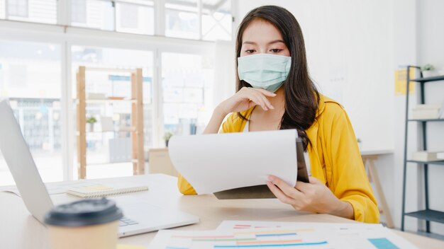 Unternehmerin aus Asien, die eine medizinische Gesichtsmaske für soziale Distanzierung in einer neuen normalen Situation zur Virenprävention trägt, während sie den Laptop wieder bei der Arbeit im Büro verwendet. Lebensstil nach dem Koronavirus.