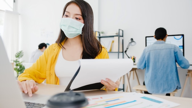 Unternehmerin aus Asien, die eine medizinische Gesichtsmaske für soziale Distanzierung in einer neuen normalen Situation zur Virenprävention trägt, während sie den Laptop wieder bei der Arbeit im Büro verwendet. Lebensstil nach dem Koronavirus.