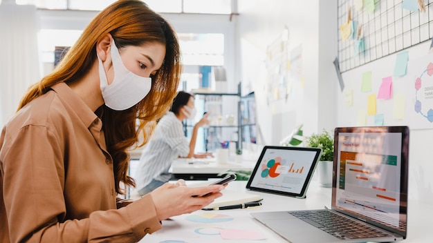 Unternehmerin aus Asien, die Gesichtsmaske zur sozialen Distanzierung in einer neuen normalen Situation zur Virenprävention trägt, während sie Laptop und Telefon bei der Arbeit im Büro verwendet.