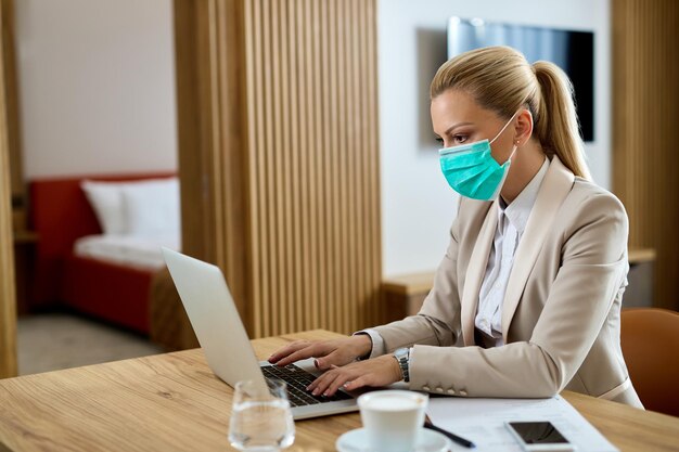 Unternehmerin mit schützender Gesichtsmaske, die eine E-Mail auf einem Computer im Hotelzimmer eingibt