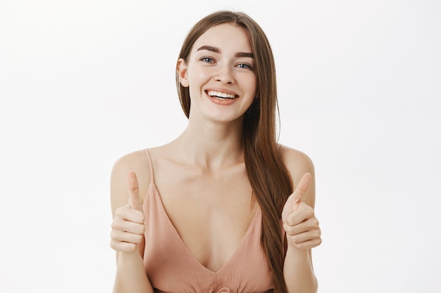 Unterstützende charmante und elegante Frau mit langen braunen Haaren im beige Kleid, das Daumen hoch zeigt