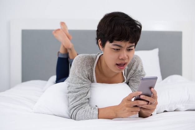 Unverschämtes asiatisches Mädchen mit Smartphone auf Bett