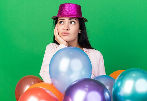 Unzufrieden aussehendes junges schönes Mädchen mit Partyhut, das hinter Ballons steht und die Hand auf die Wange legt, isoliert auf grüner Wand