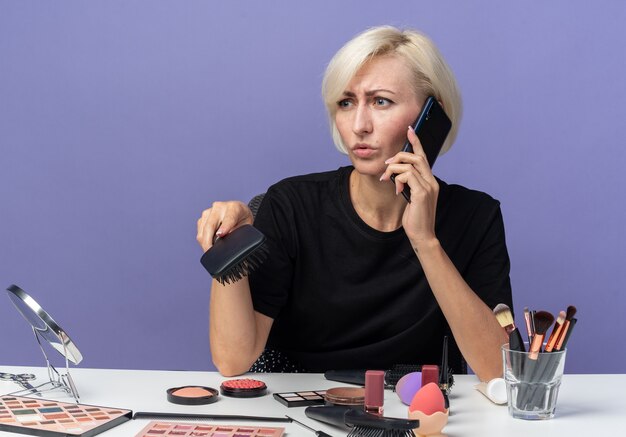 Unzufrieden aussehendes junges schönes Mädchen sitzt am Tisch mit Make-up-Tools spricht am Telefon und hält Kamm isoliert auf blauem Hintergrund