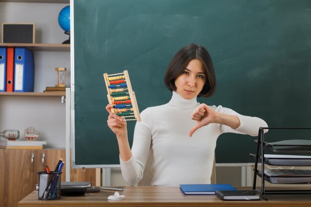 unzufrieden mit Daumen nach unten junge Lehrerin mit Abakus am Schreibtisch sitzend mit Schulwerkzeugen im Klassenzimmer